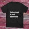 Black T-shirt with the phrase "Fatherhood, A Reel Adventure" in bold white text, shown against a light blue background.