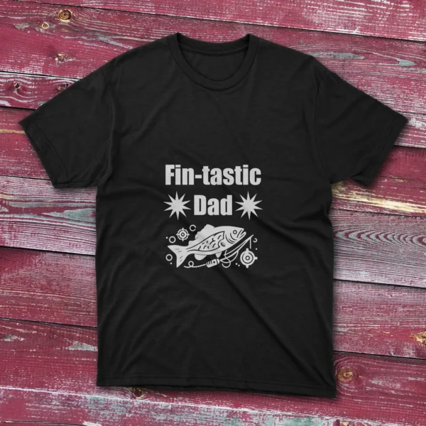 Black T-shirt on a light red wooden ground featuring "Fin-Tastic Dad" with two stars and a decorative fish illustration in white.