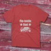 red T-shirt on a light red wooden ground featuring "Fin-Tastic Dad" with two stars and a decorative fish illustration in white.