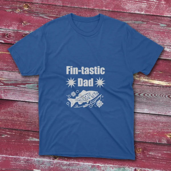 royal blue T-shirt on a light red wooden ground featuring "Fin-Tastic Dad" with two stars and a decorative fish illustration in white.