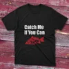 Black fishing T-shirt featuring the phrase "Catch me if you can" in bold white text with a red fish illustration, set against a light blue background.